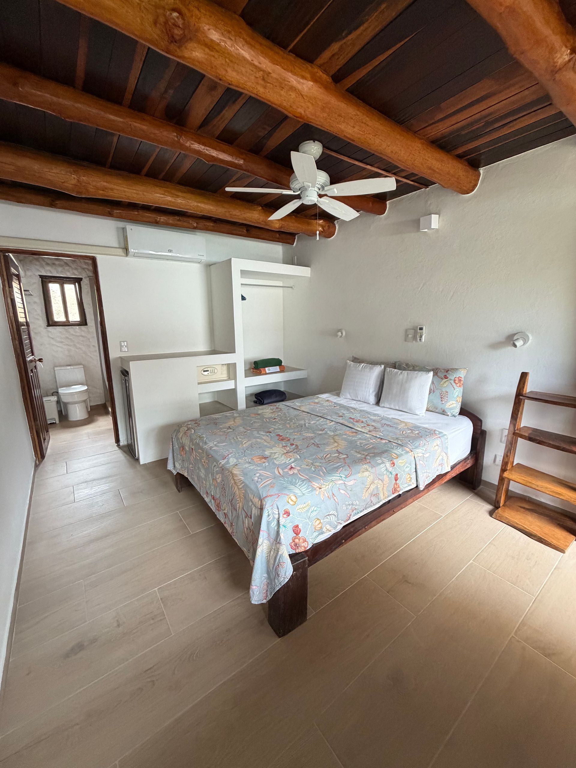 Bedroom with exposed wooden ceiling, ceiling fan, bed, and small bathroom visible through an open doorway.