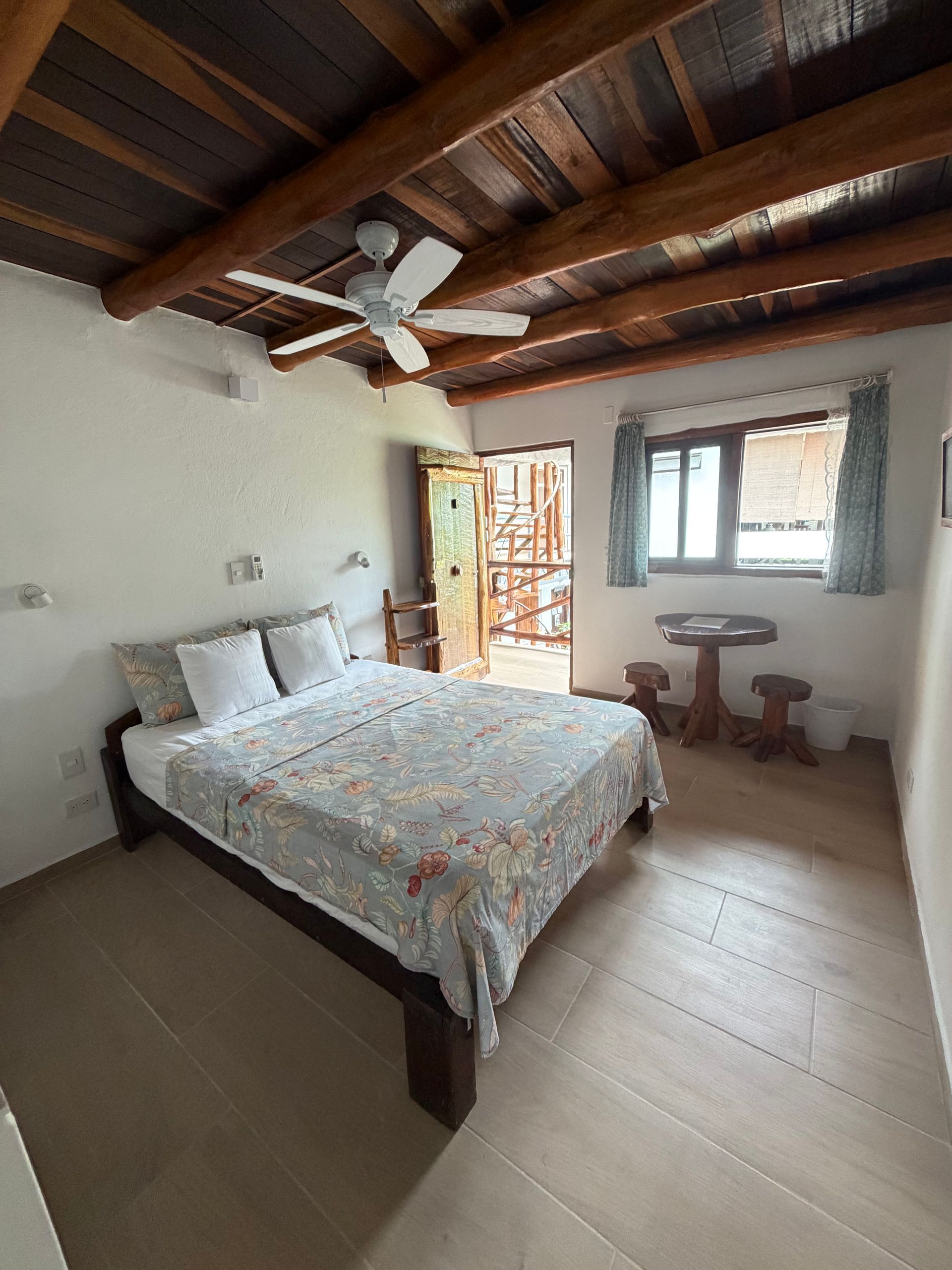 Rustic bedroom with bed, ceiling fan, wooden beams, and window with table and chairs