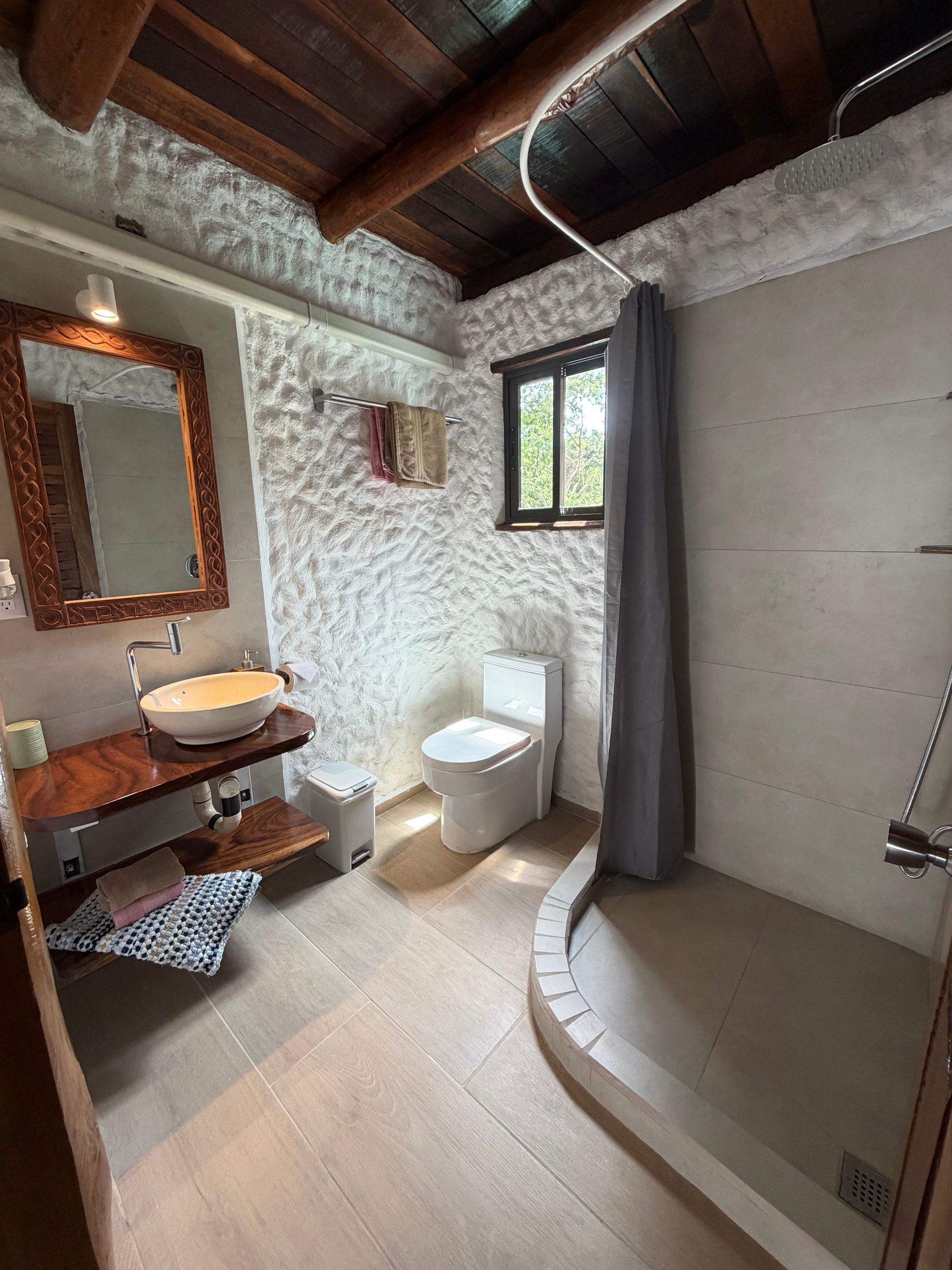Rustic bathroom with stone walls, glass shower, white toilet, wooden sink, and small window.