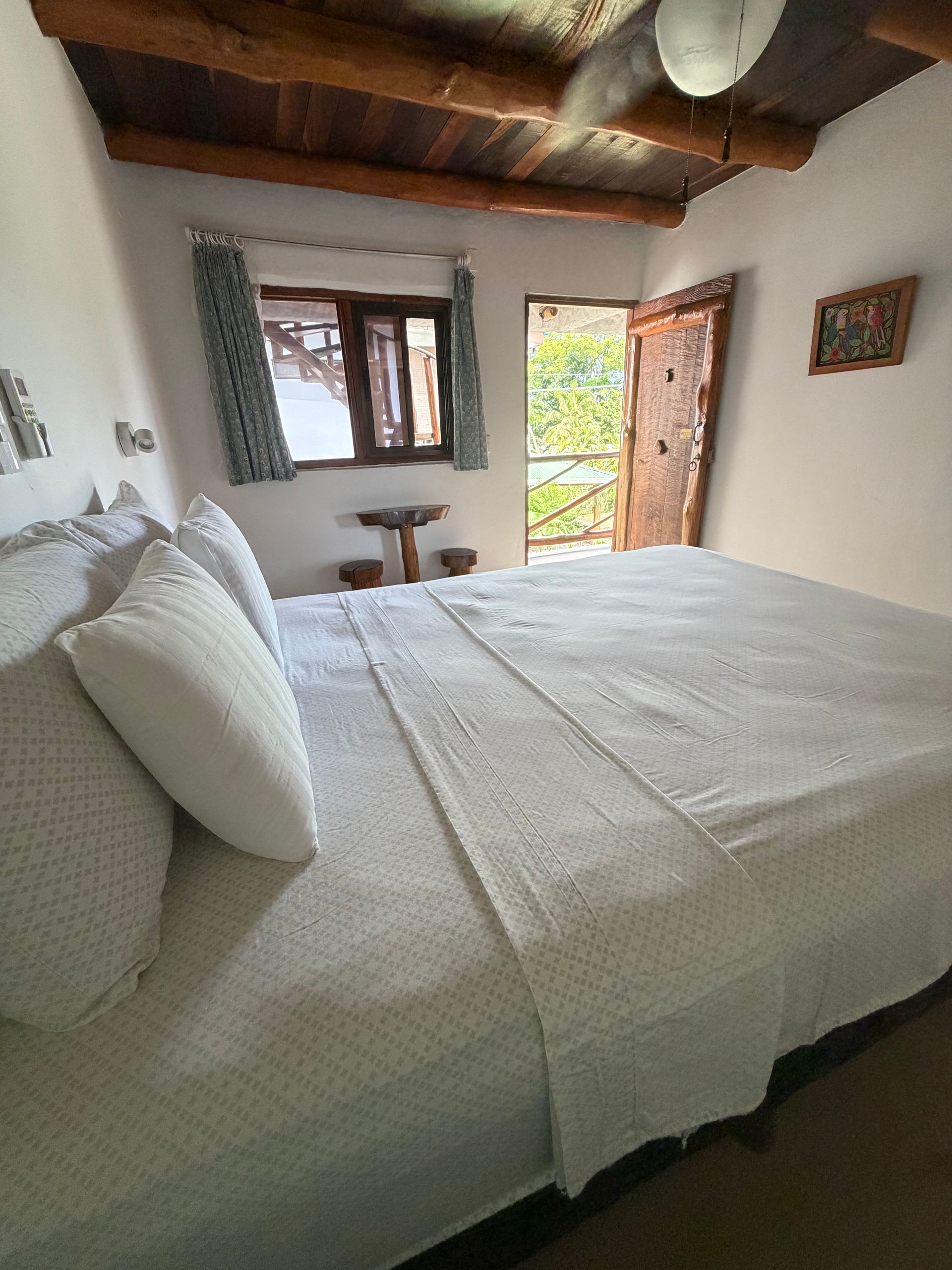 White bed in a rustic bedroom with wooden ceiling, window, open door, and daylight outside