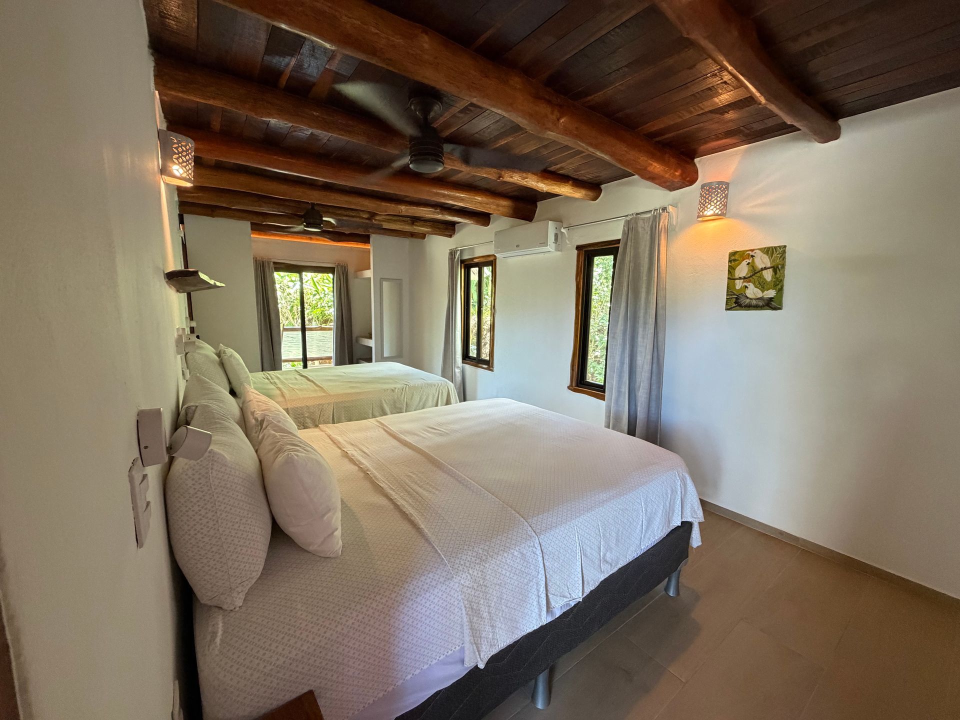 Bright bedroom with two white beds, exposed wood ceiling, and windows overlooking greenery