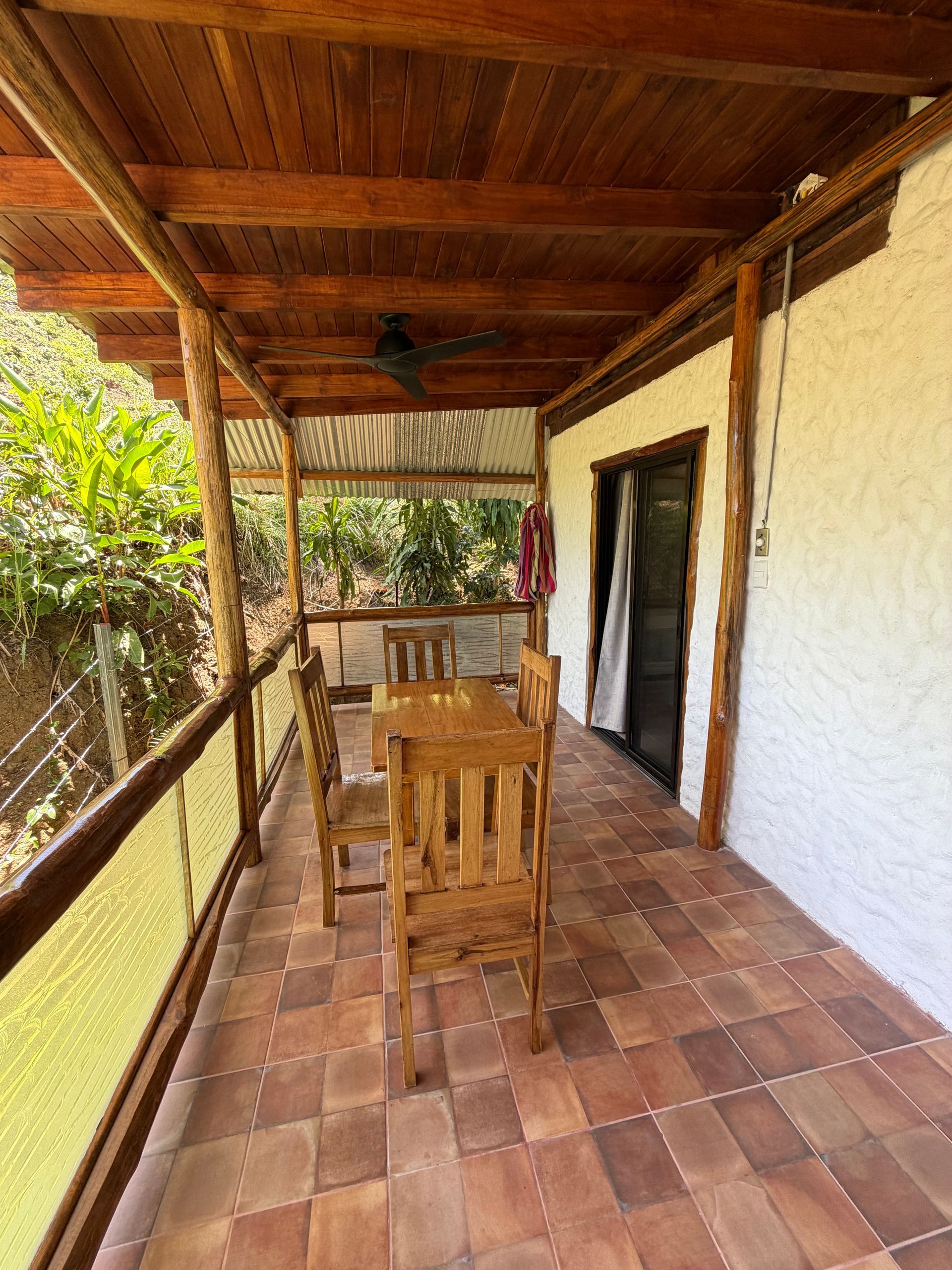 Covered patio with wooden table and chairs, terracotta tile floor, and open doorway beside a white stucco wall