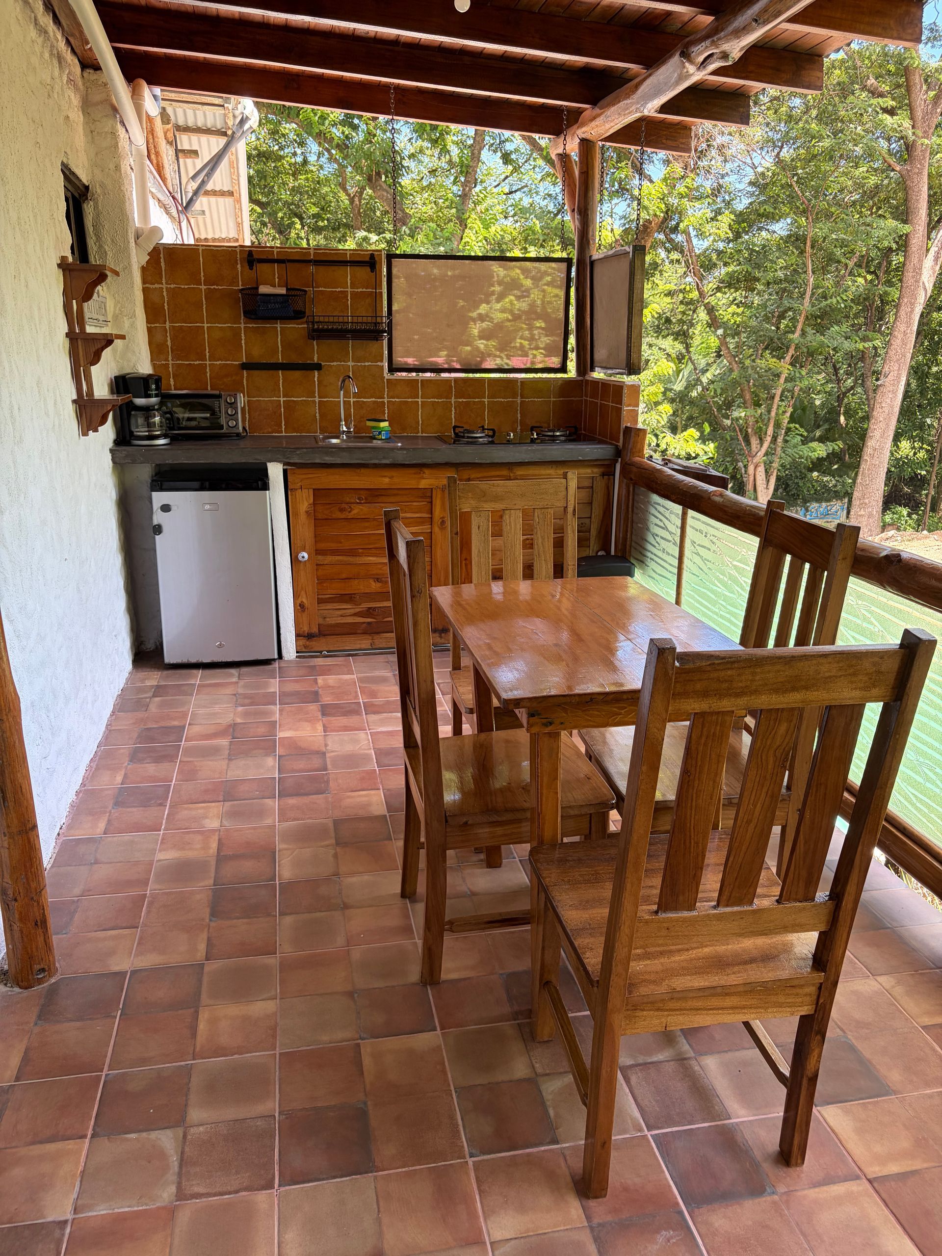 Covered outdoor kitchen with wooden table and chairs, brick floor, and sink under a tiled roof.