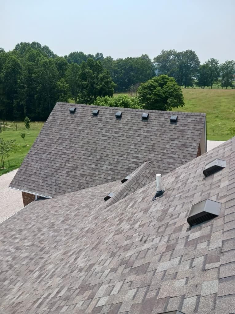 Roofs of a house with asphalt shingles, vents, and a tree-lined hillside in the background.