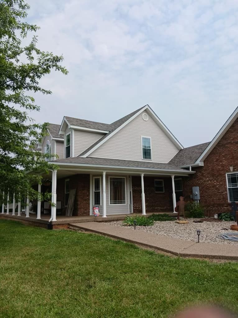 Two-story house with a large porch and brick accents, surrounded by greenery under a cloudy sky.