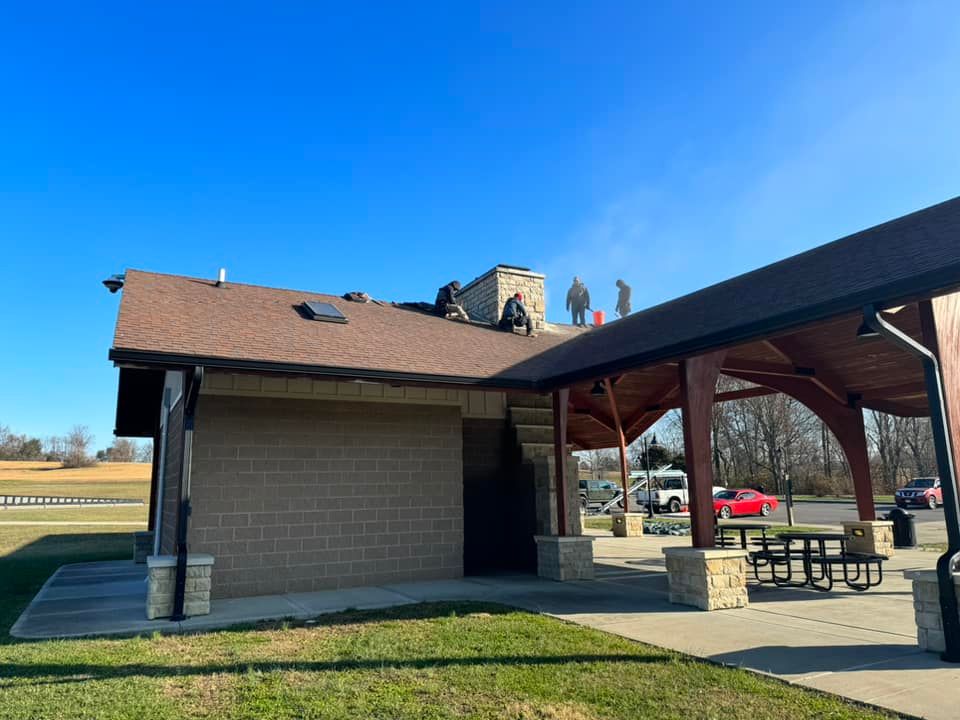 Workers on a brown shingled roof of a public restroom with an attached picnic area under a clear, blue sky.