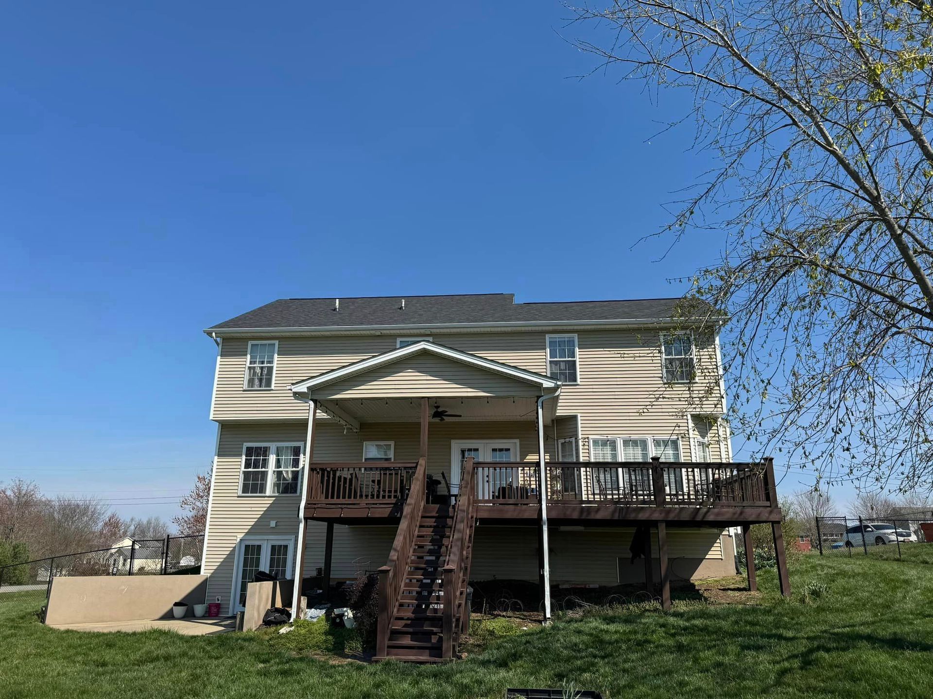 Back of a two-story beige house with a dark wooden deck and stairs, on a grassy hill under a blue sky.