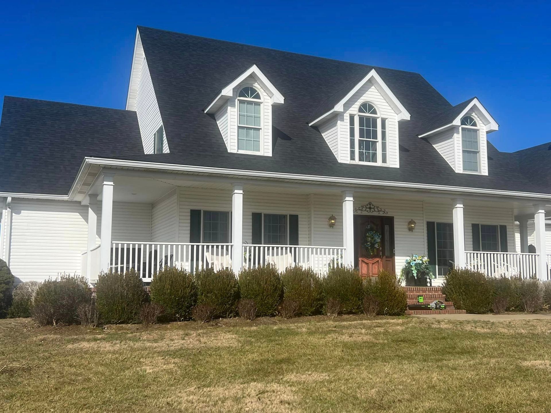 White house with black roof, covered porch, and three dormer windows under a clear blue sky.