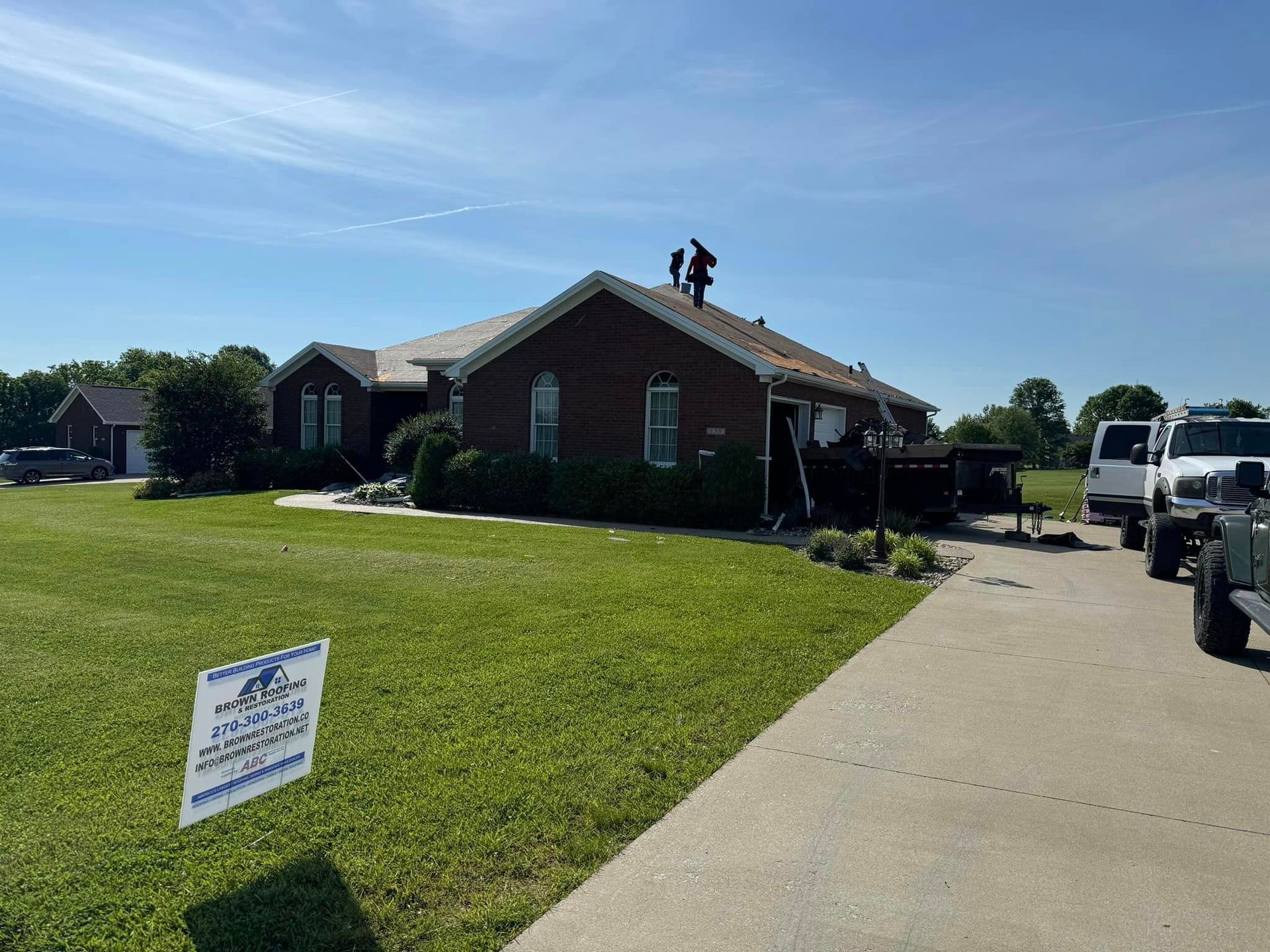 Roofers working on a house with brown siding, a sign in the yard, and vehicles parked nearby on a sunny day.
