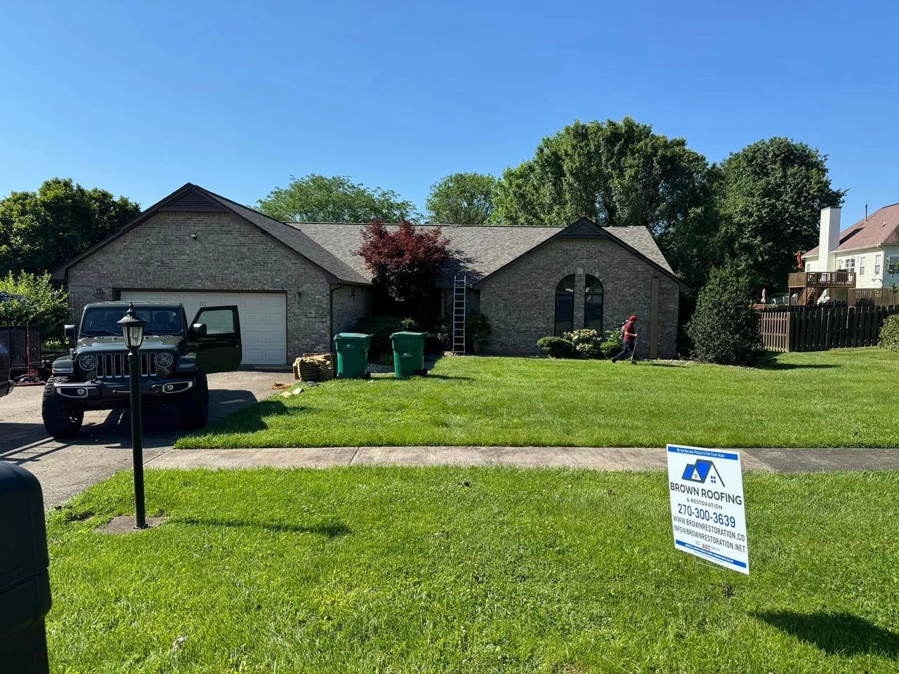 House with brick facade, green lawn, and a Jeep parked in the driveway. A ladder leans against the roof.