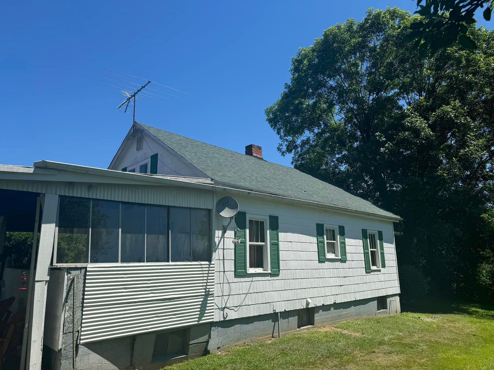 White house with green shutters, metal roof, antenna, and enclosed porch under a blue sky.