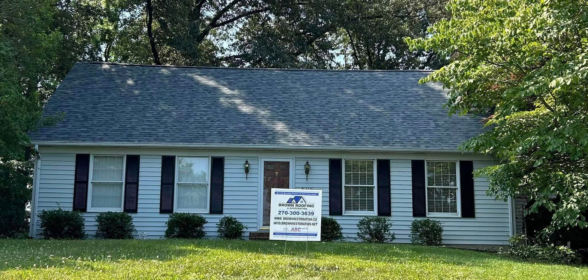 A small, light-blue house with black shutters, a dark roof, and a lawn. A sign is in front.