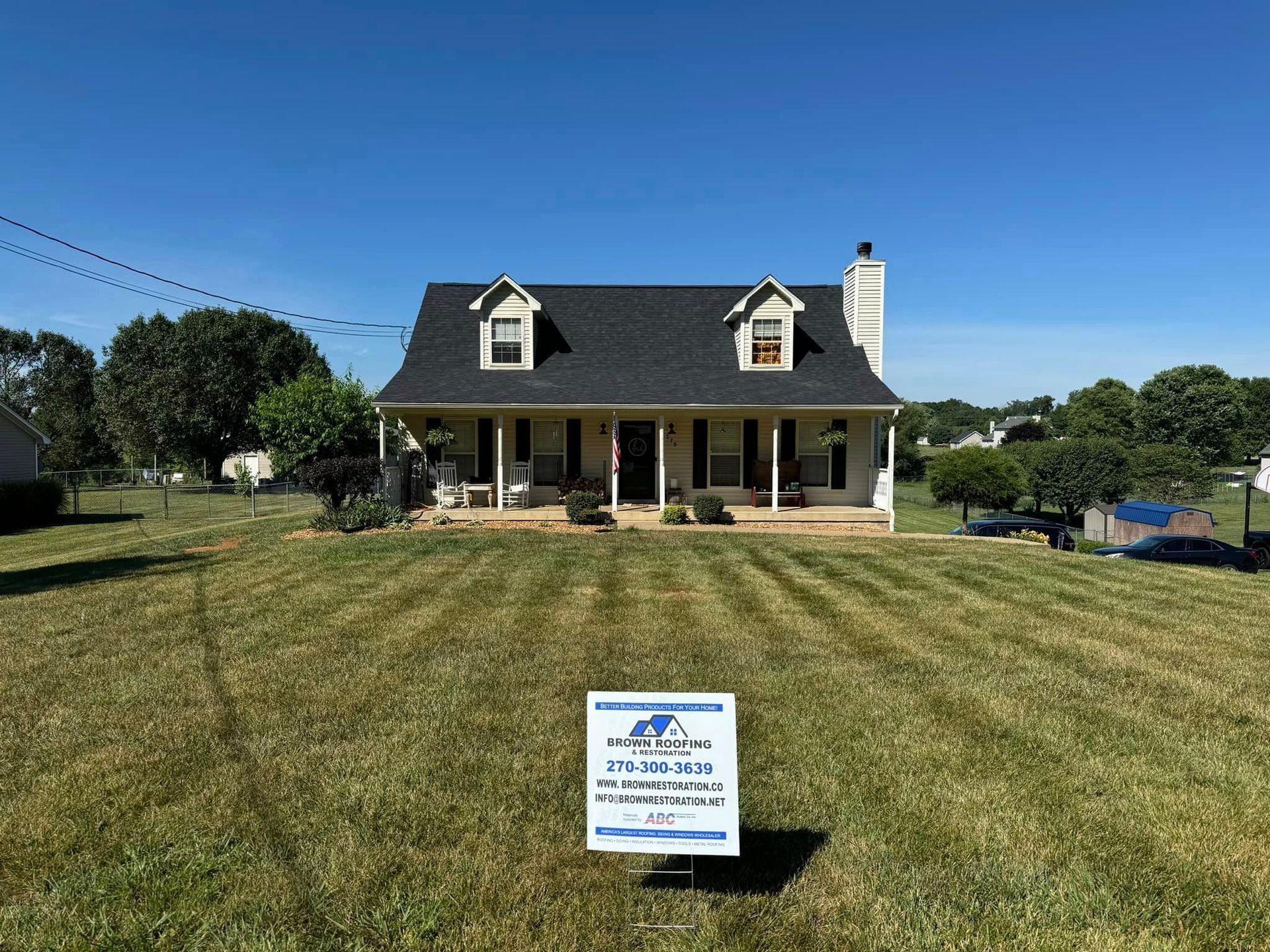 House with dark roof and dormers on a grassy lot with a sign in front, under a blue sky.