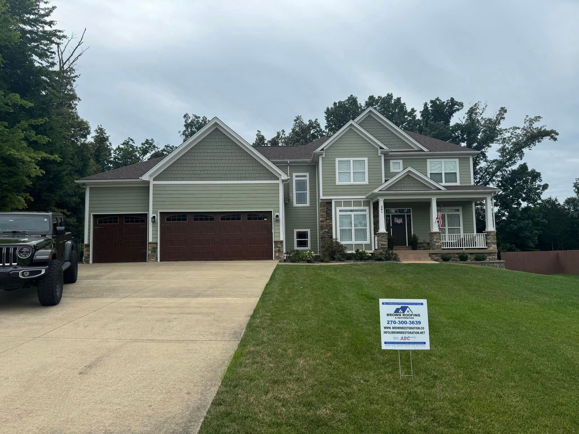 Green house with brown garage doors and a Jeep parked in the driveway. Sign in yard.