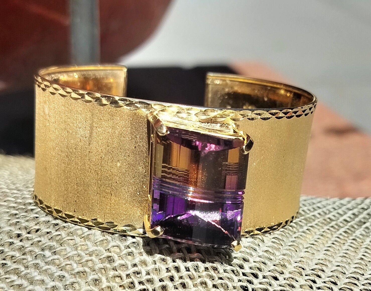 Gold cuff bracelet with a purple rectangular gemstone centerpiece on a textured surface