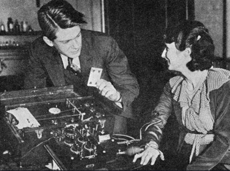 A man demonstrates card trick for a woman. He holds cards over a box with dials. Both smile.
