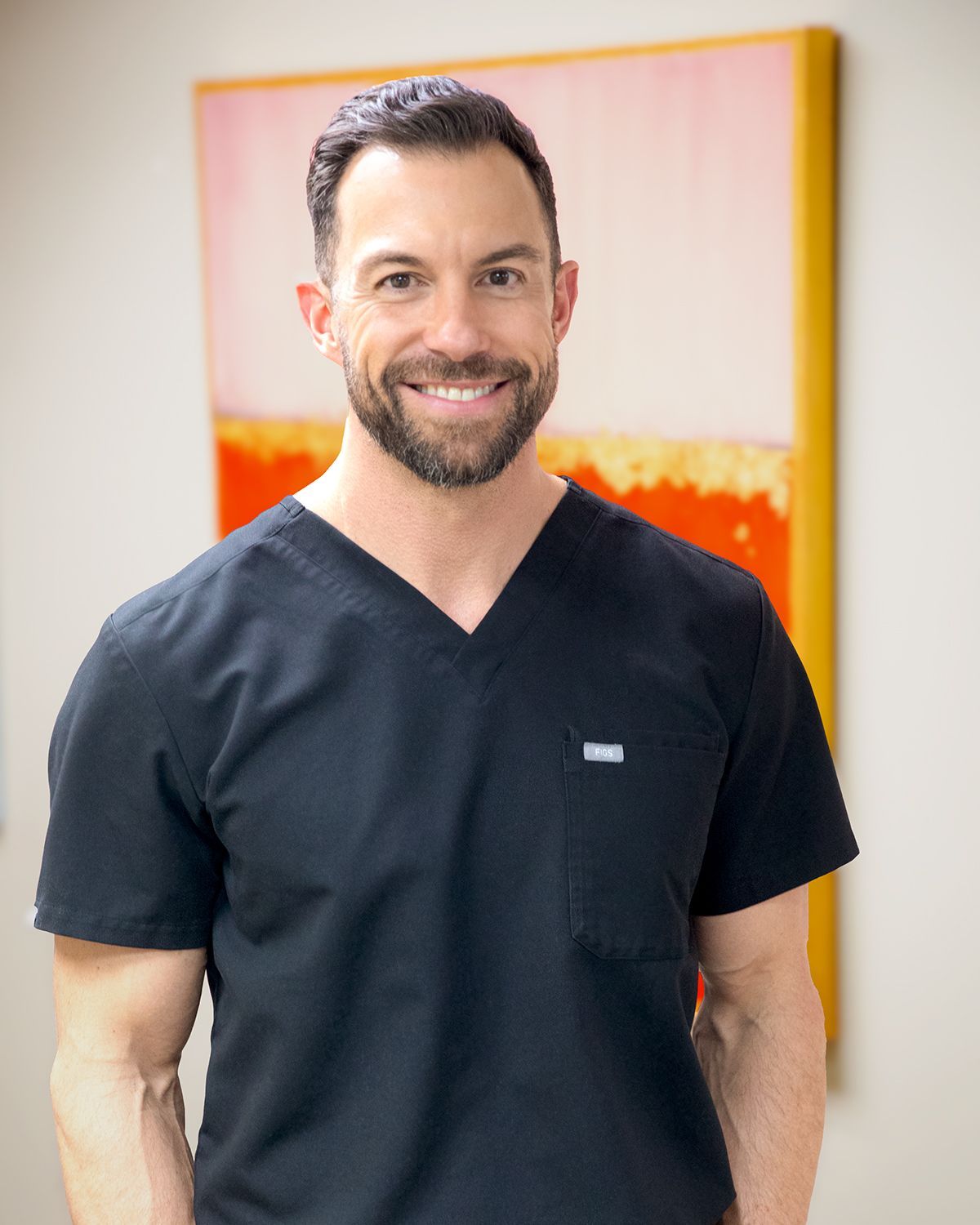A man in a black scrub top is smiling in front of a painting.