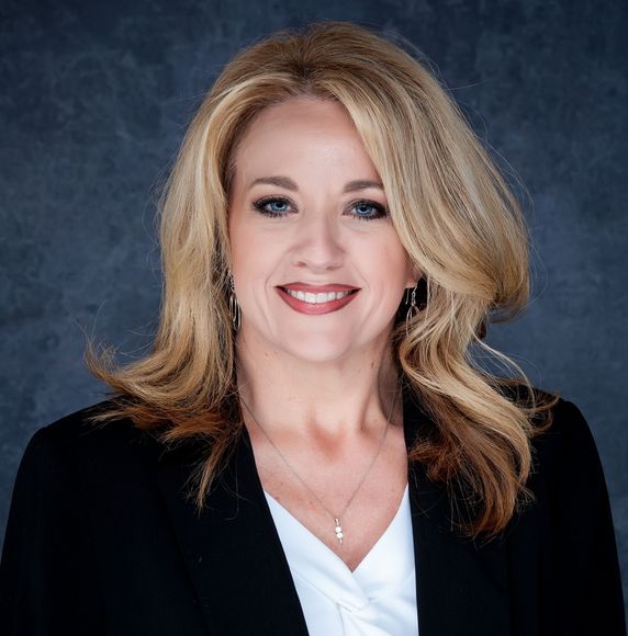Studio headshot of a smiling woman in a black blazer and white blouse on a blue-gray backdrop
