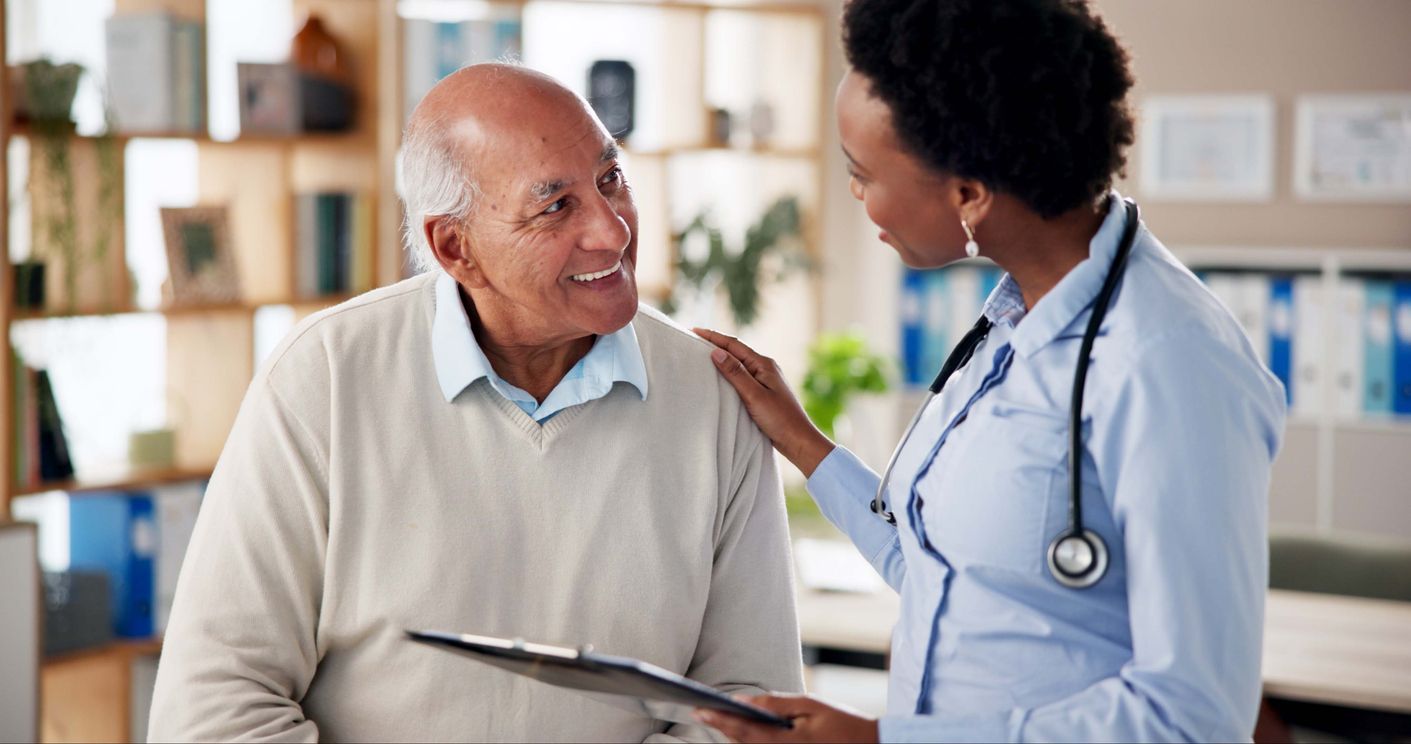 Doctor speaking with a patient in a bright clinic, holding a clipboard and resting a hand on the patient’s shoulder