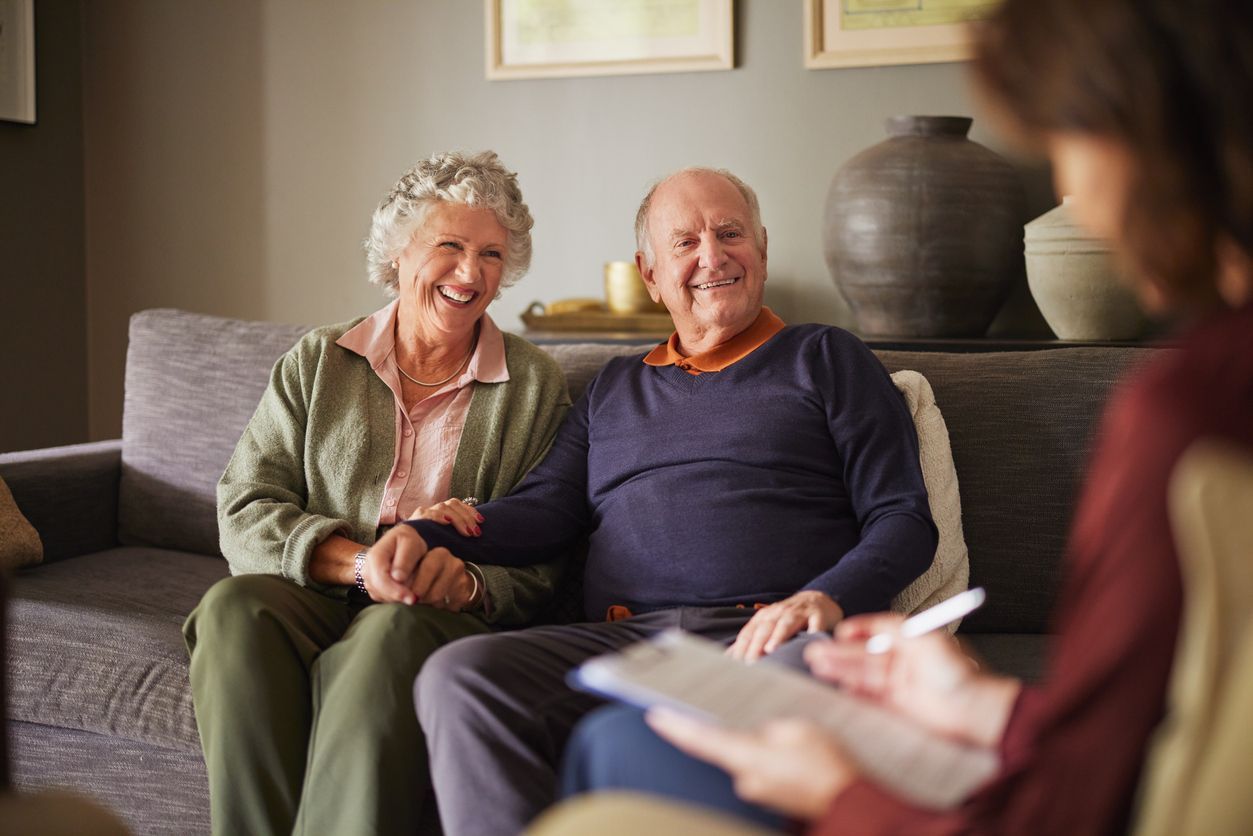 Elderly couple smiling, holding hands on couch, meeting with a professional.