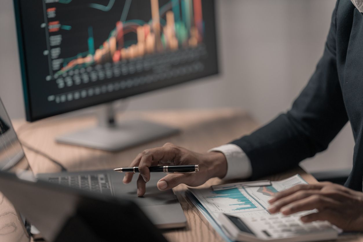 Person reviewing financial charts on a laptop at a desk with documents and a monitor displaying data.