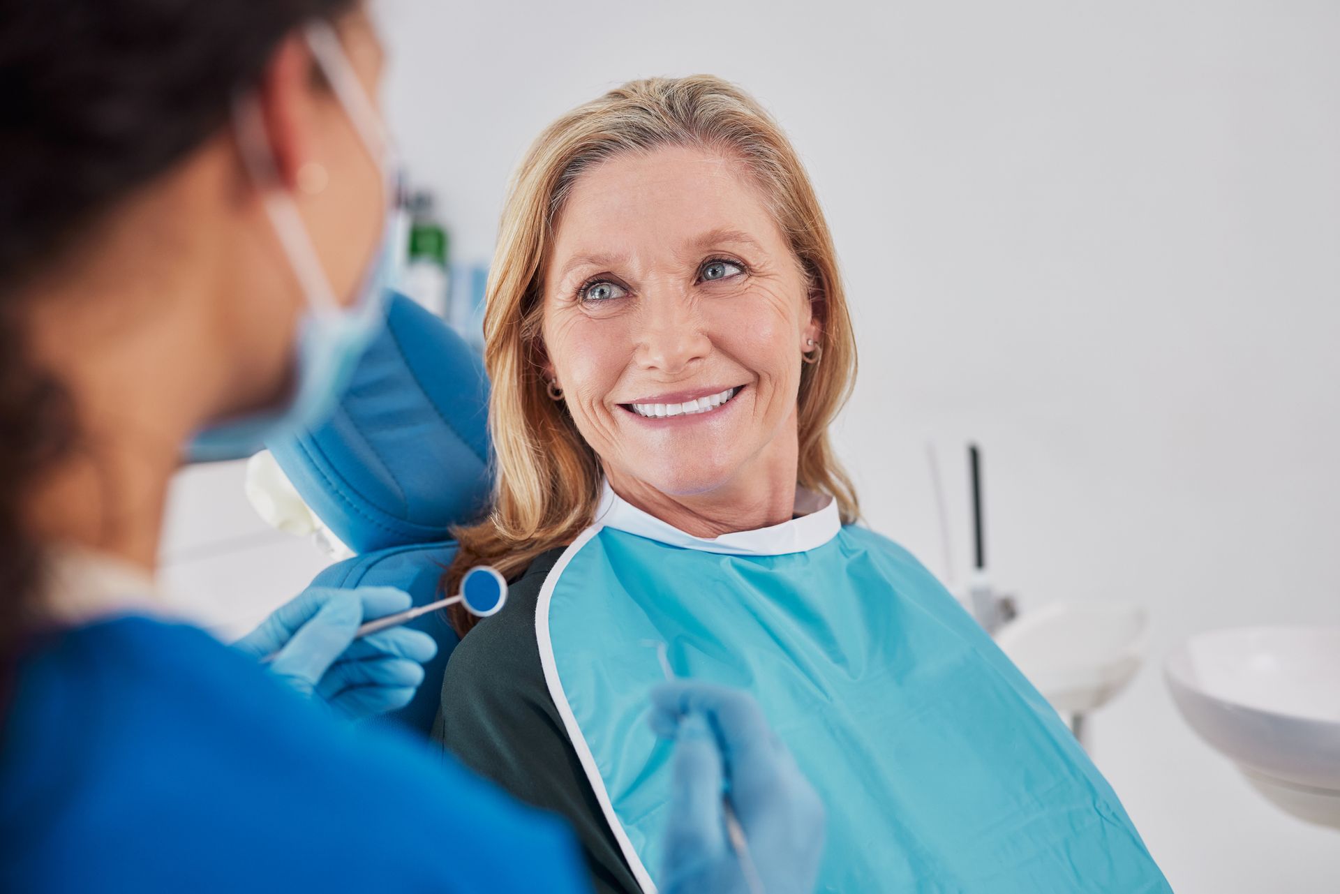 Patient smiling in dental chair while dentist prepares treatment, with blue bib and mask in a bright clinic.