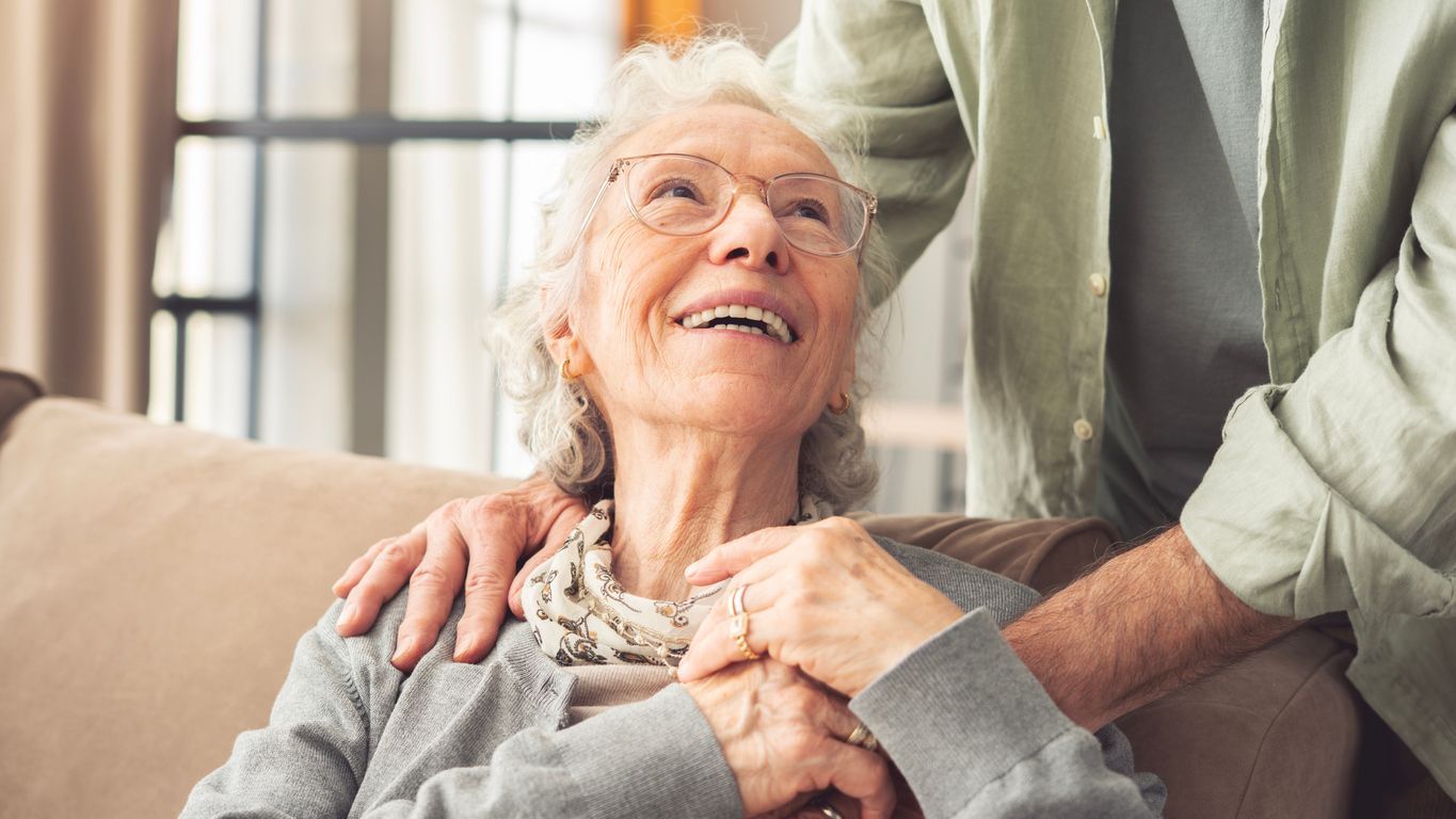 Smiling older woman reclining on a sofa while someone gently holds her hand and shoulder
