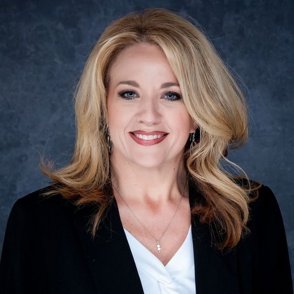 Woman with blonde hair wearing a black blazer smiles, white shirt, against a dark blue background.