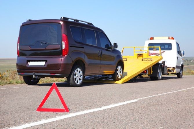 Tow truck loading a dark minivan on a roadside, with a red warning triangle in the foreground.