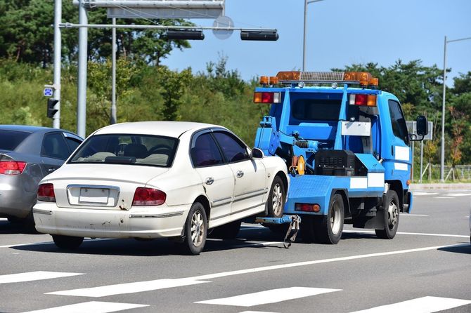 White sedan being towed by a blue tow truck at an intersection in daylight.