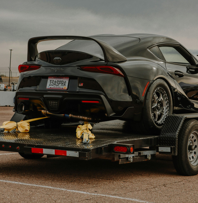 Black sports car on a trailer with a large rear wing, parked on a road