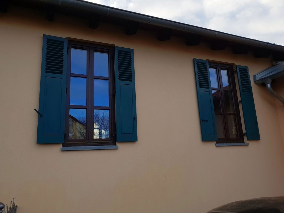 Due finestre con persiane color verde acqua su un edificio chiaro, che riflettono il cielo e l'ambiente circostante.