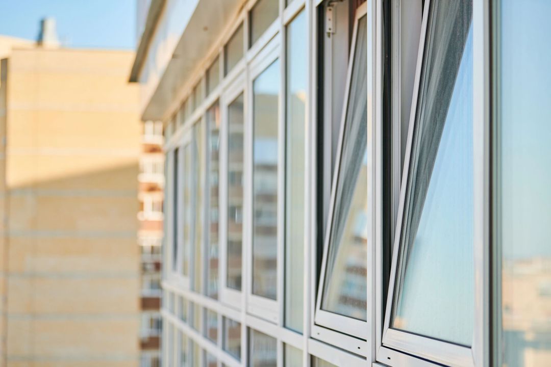 Finestre con cornici bianche sulla facciata di un edificio. Una finestra parzialmente aperta, con una zanzariera. Cielo azzurro e limpido riflesso.