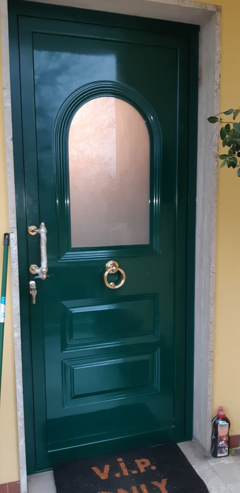 Un portone d'ingresso verde con una finestra ad arco e un batacchio dorato. Davanti c'è uno zerbino con la scritta