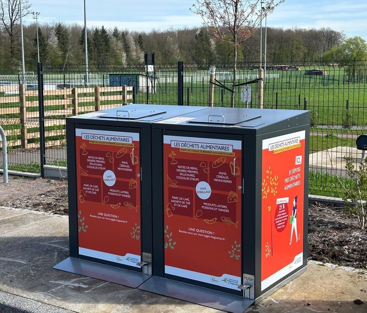 Deux conteneurs de recyclage gris foncé avec des graphismes promotionnels rouges et blancs, à l'extérieur.