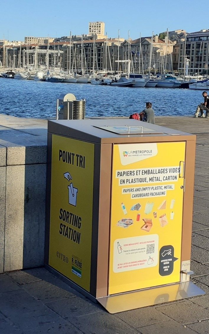 Station de tri des déchets jaune près de l'eau, avec la ville en arrière-plan.