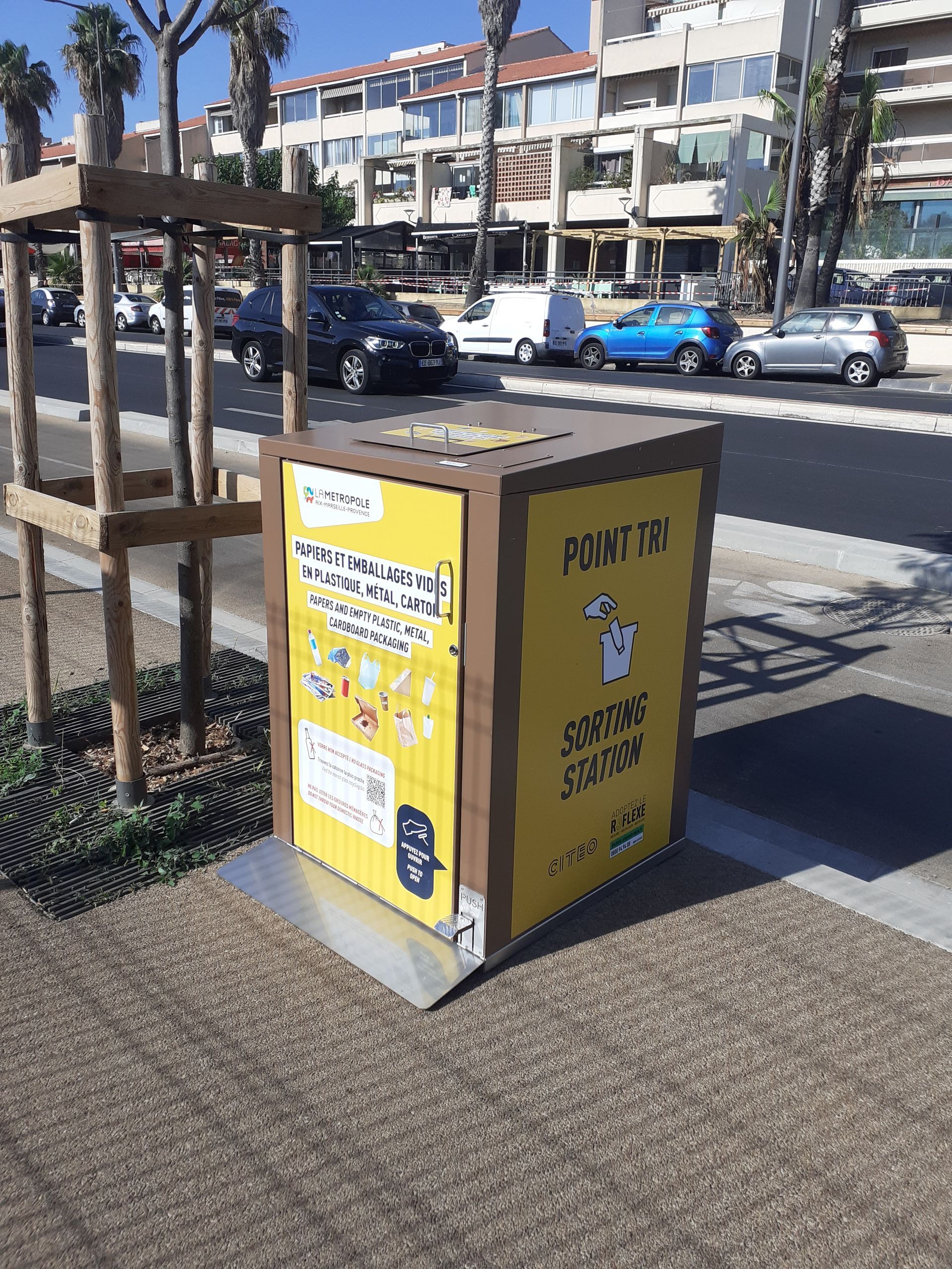 Poubelles de recyclage jaunes et marron sur un trottoir, devant des immeubles et des voitures, par une journée ensoleillée.