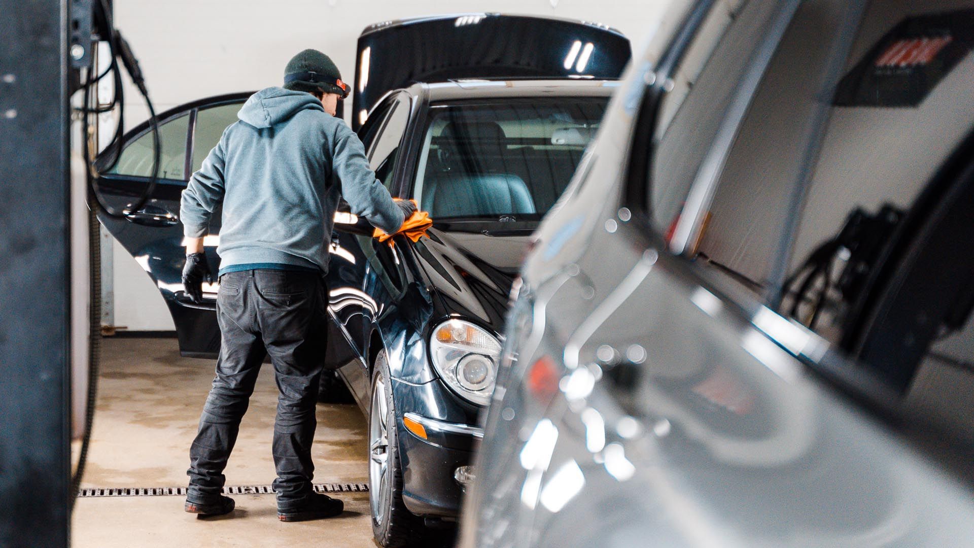 Person cleaning a black car in a garage; wearing a gray hoodie and black gloves.