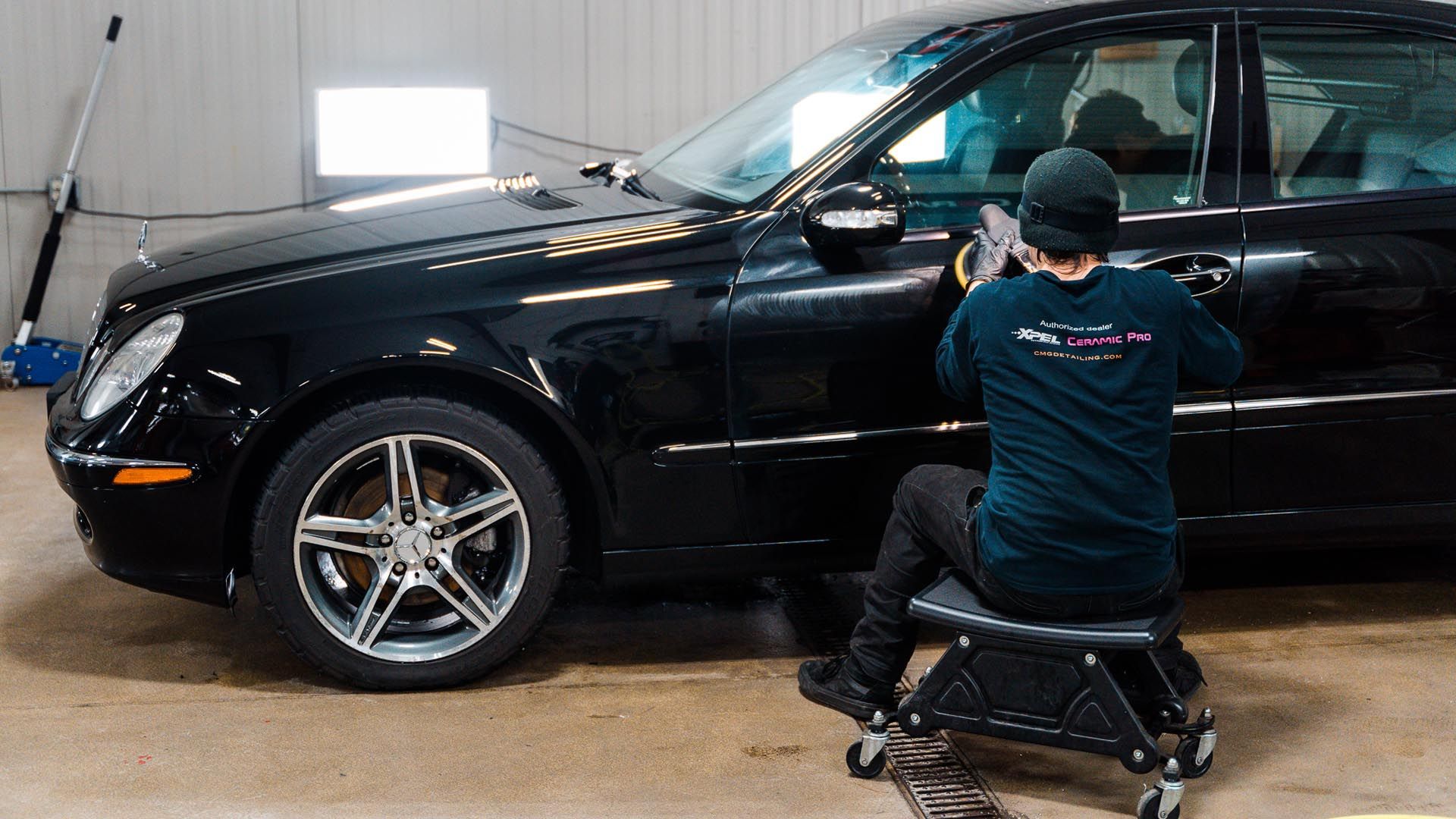 Person polishing a black car door, sitting on a rolling stool indoors.