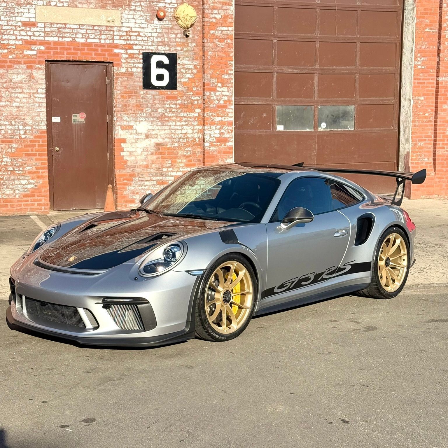 Silver Porsche 911 GT3 RS with gold wheels parked in front of a brick building with a large garage door.