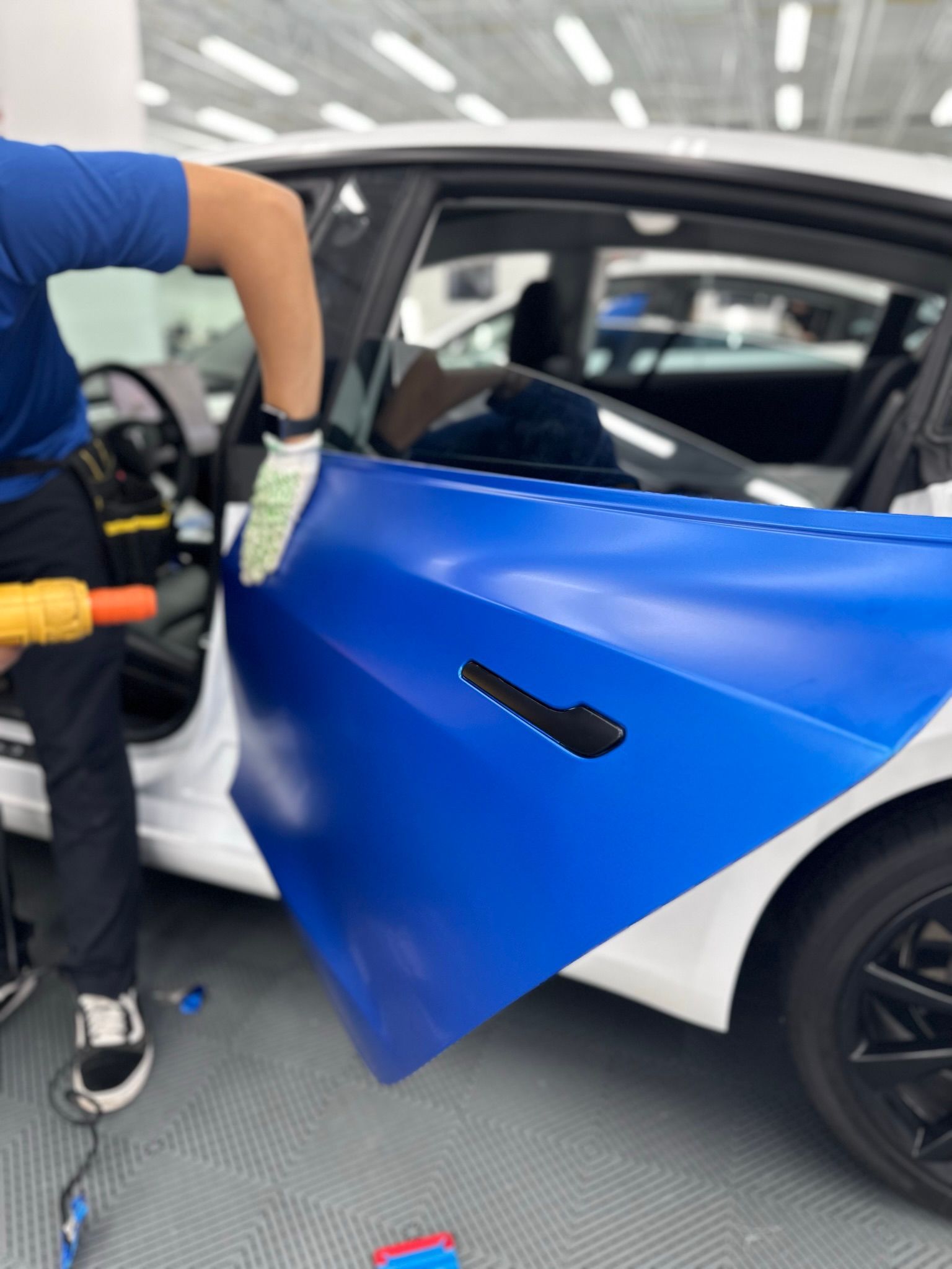 Person wrapping a blue vinyl sheet on a white car door, inside a garage.