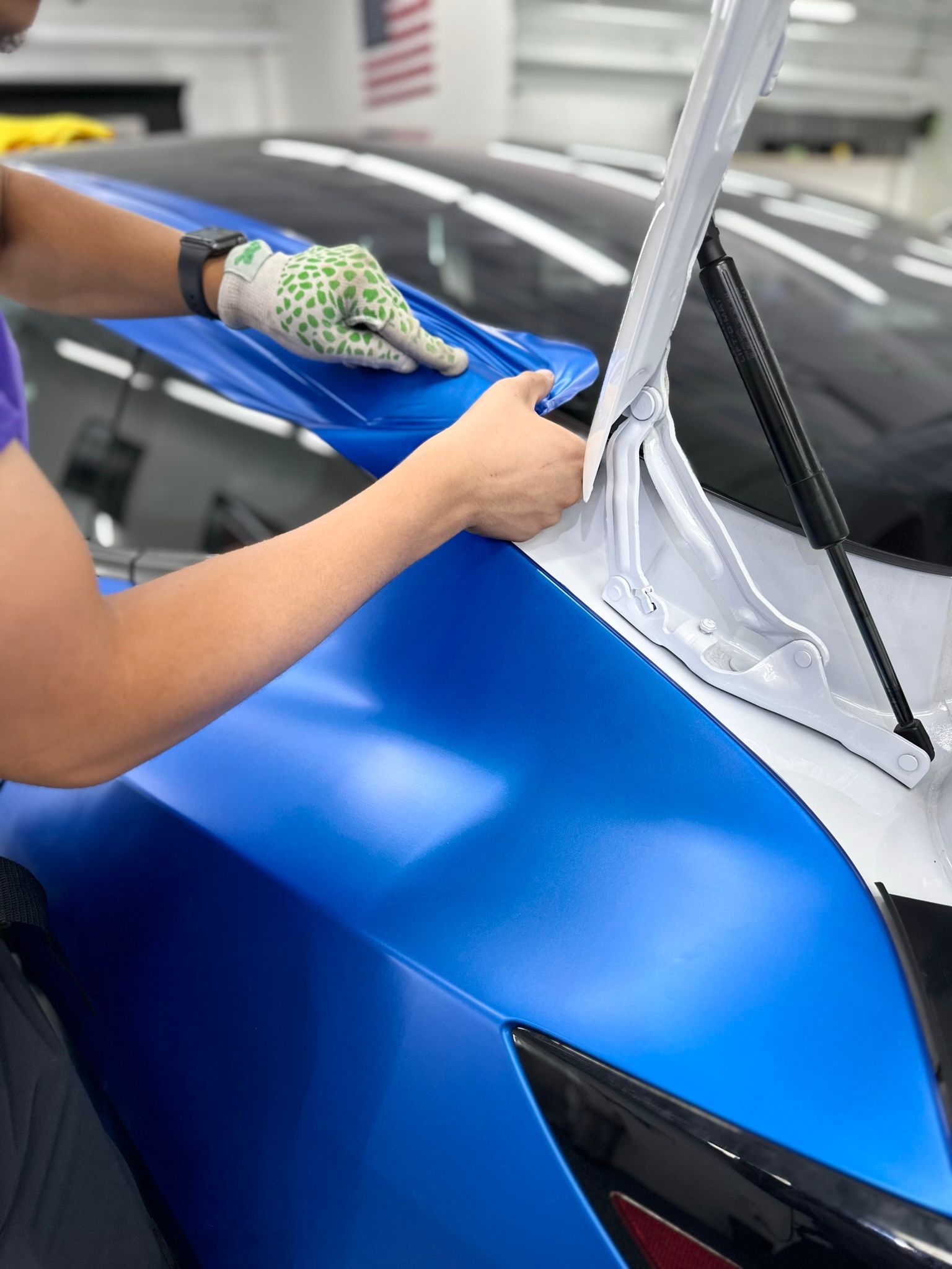 Person wrapping a blue car's hood with vinyl in a shop. They wear gloves, with an open hood.
