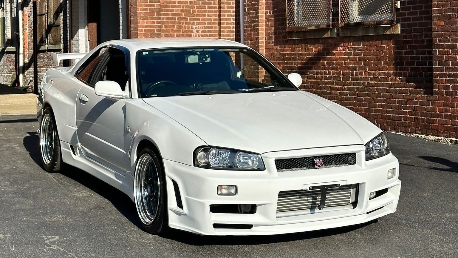 White Nissan Skyline R34 sports car parked in front of a brick building.