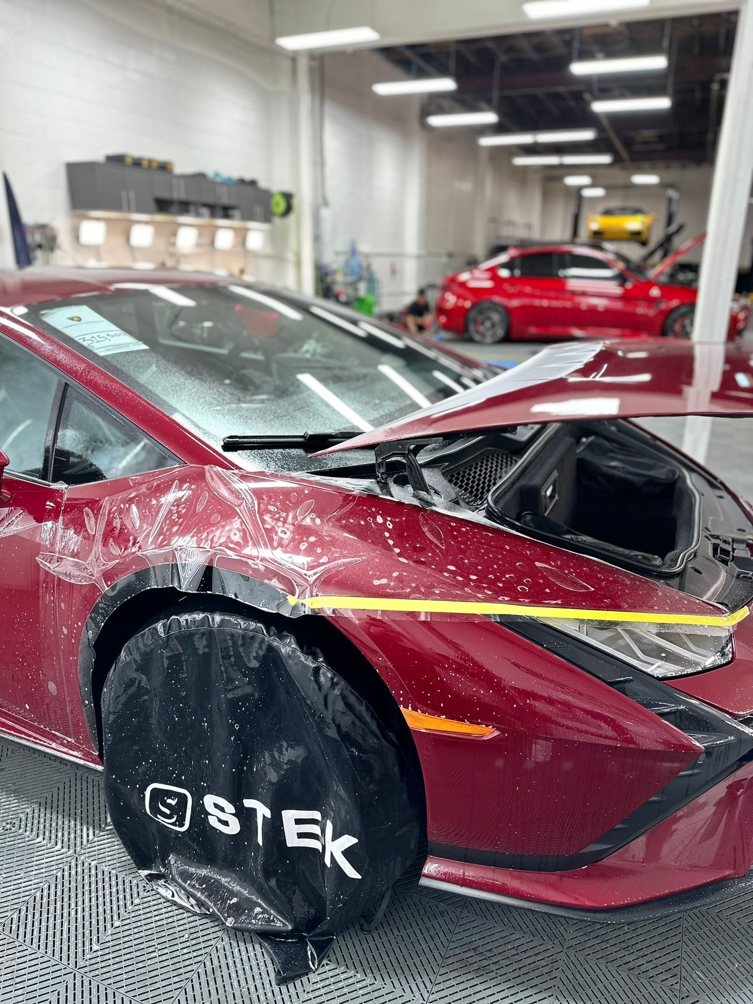 Maroon Lamborghini in a workshop, covered in water, being worked on, with STEK tire cover.