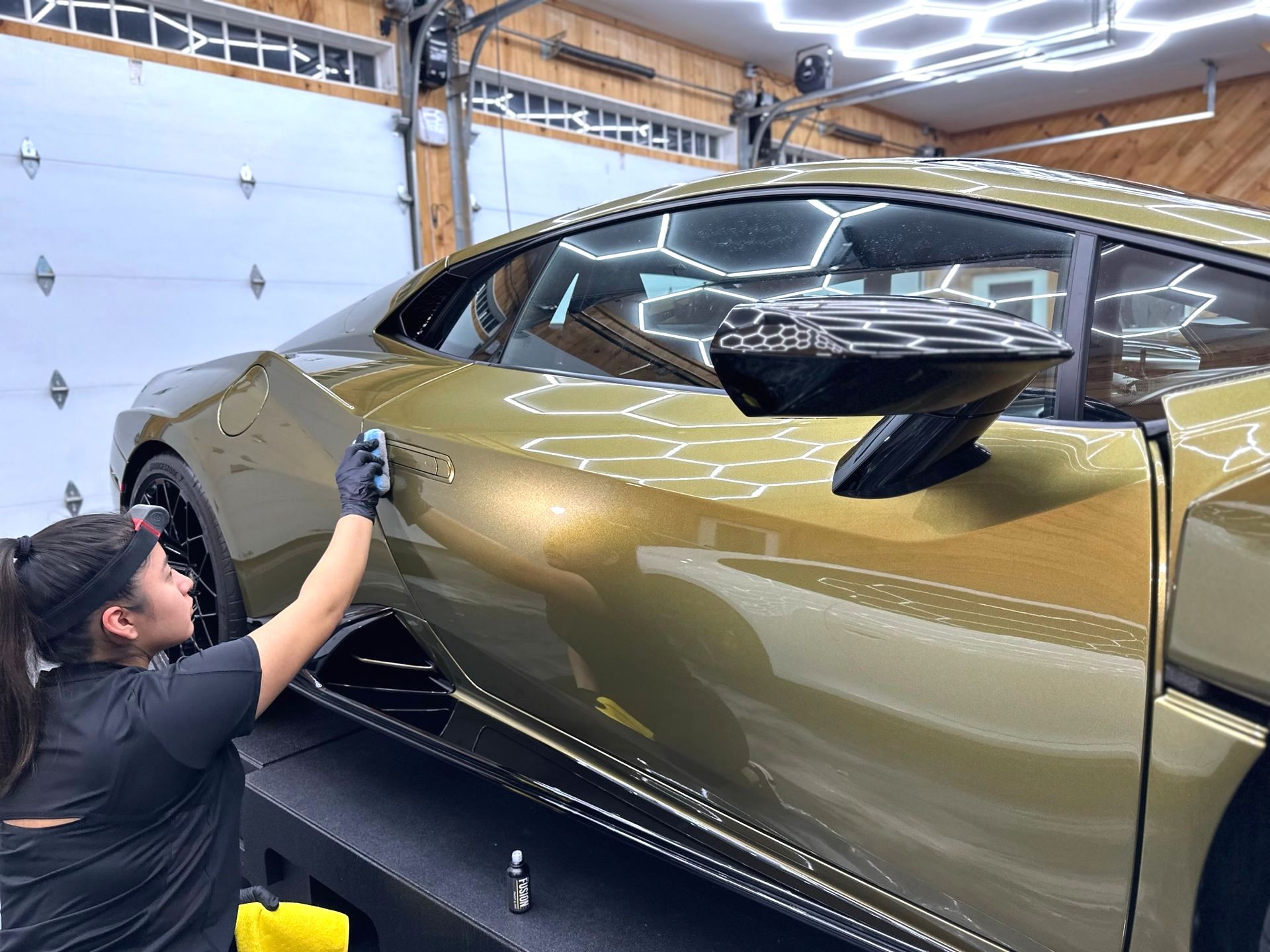 Woman polishing a gold sports car in a garage with bright lights.