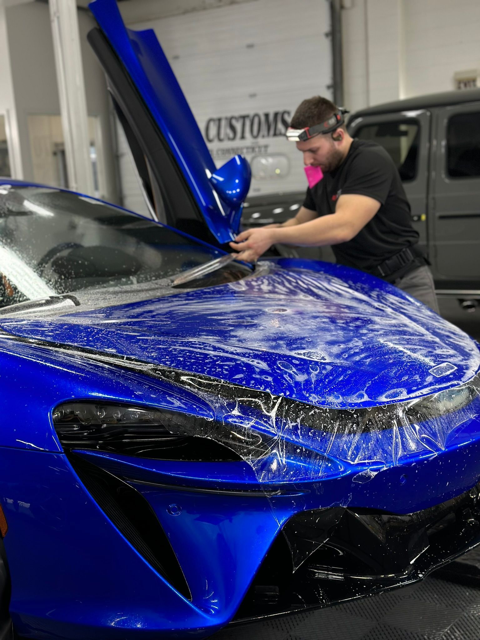 Man applying film to a blue McLaren car. The car's door is open. He works inside a shop.