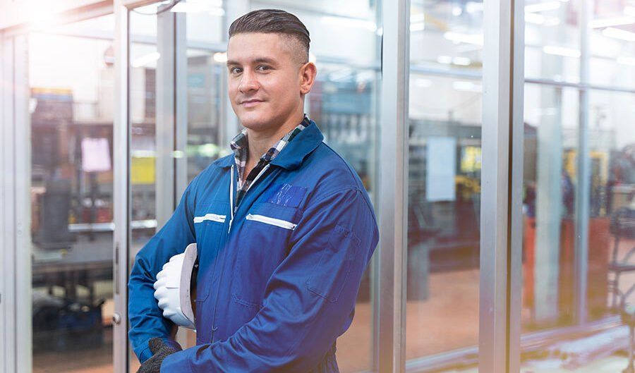 A man in a blue jumpsuit is holding a hard hat in a factory.