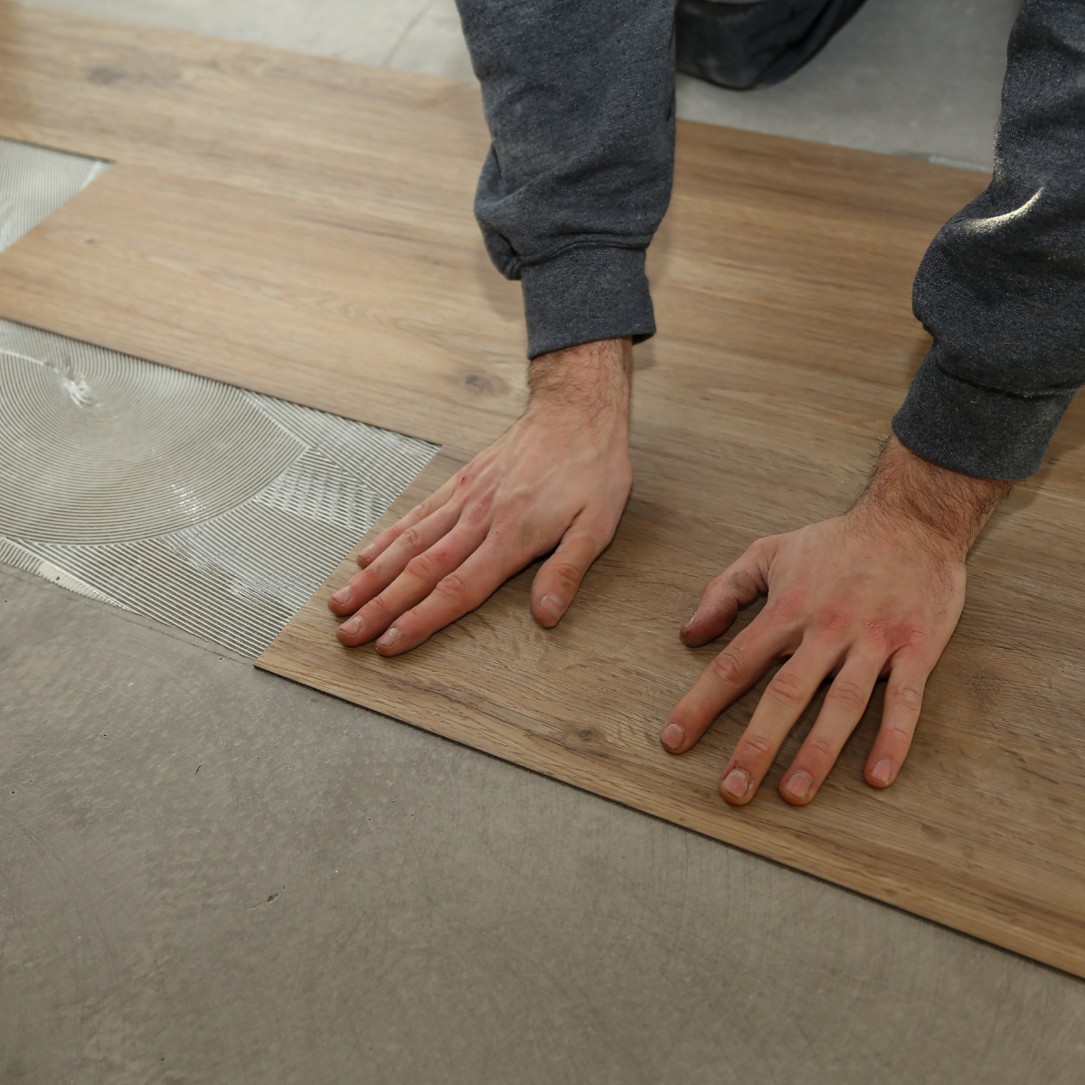 Hands aligning wood flooring planks beside a section of adhesive on a subfloor