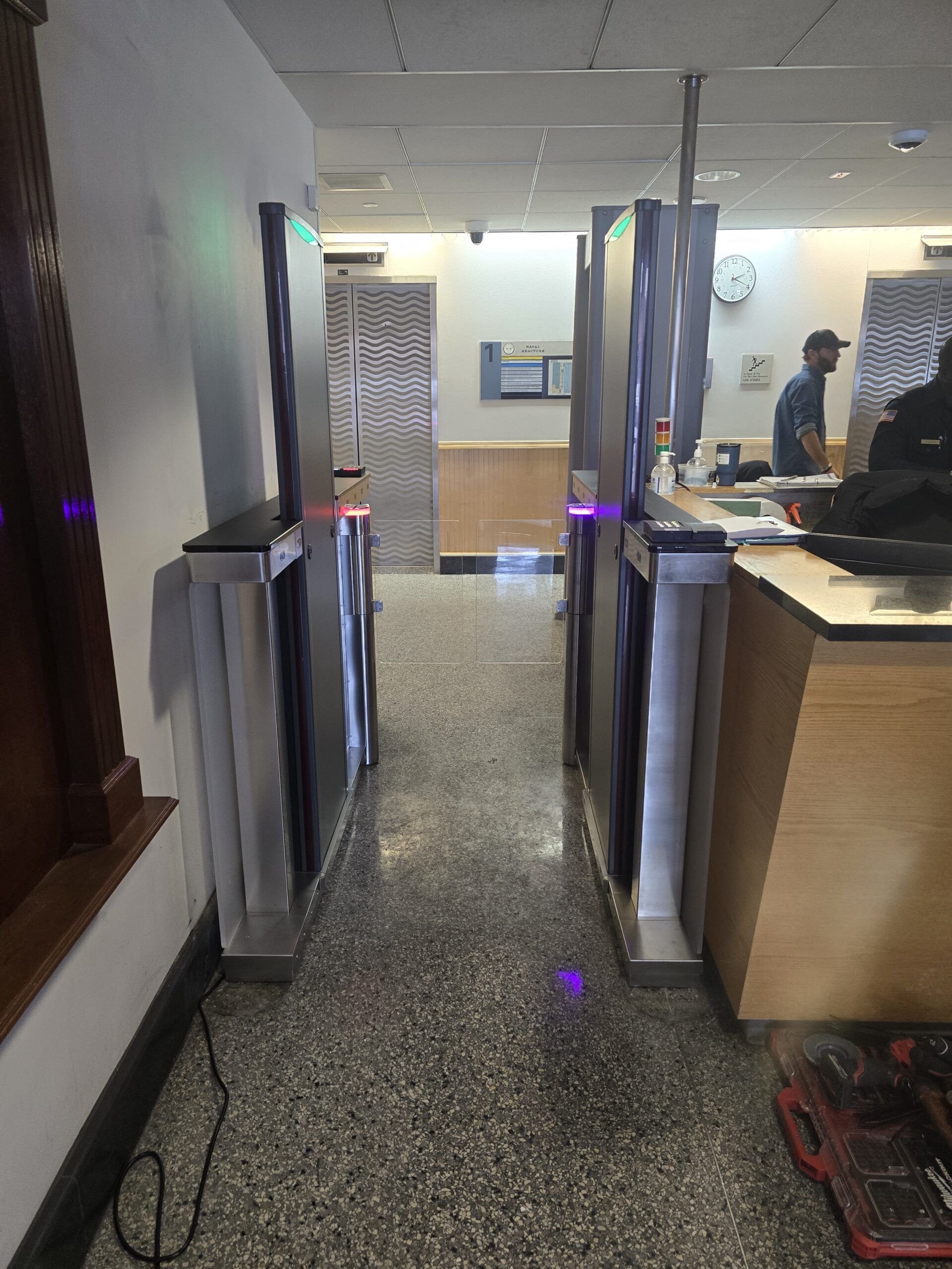 Security turnstiles in a building entrance with a person in the background. Fluorescent lights and a gray floor.