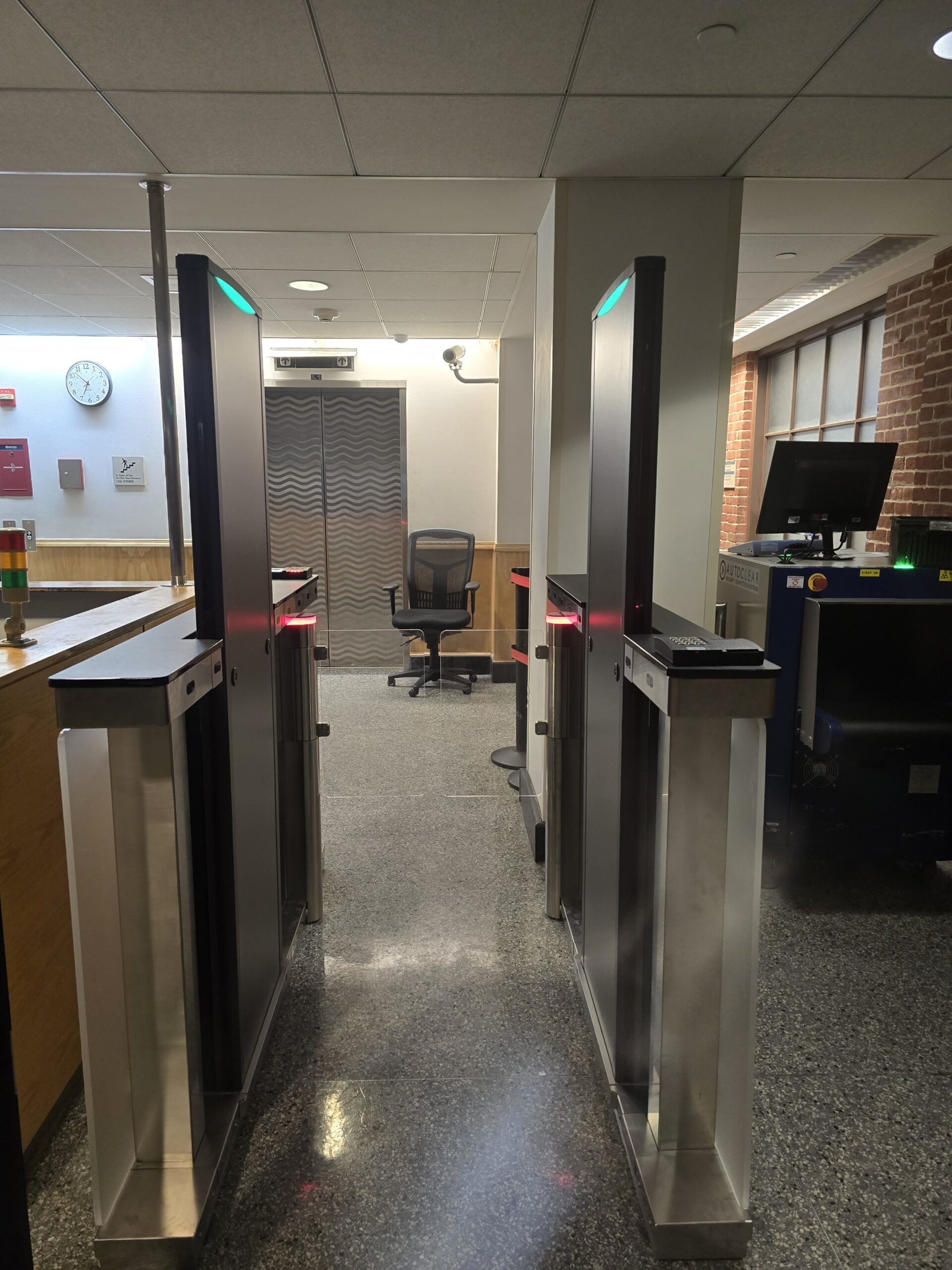 Security turnstiles with red laser lines. A door and a desk are in the background.